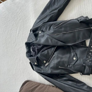 Svart skinnjacka från Zara - Snygg svart skinnjacka från Zara med coola dragkedjedetaljer och bälte i midjan. Perfekt för att ge din outfit en edgy touch. Jackan har långa ärmar, använd en gång. Passar en som har S också 