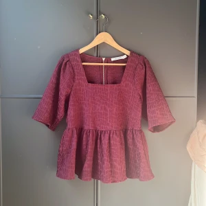 Vinröd blus från Carin Wester  - Säljer en elegant vinröd blus från Carin Wester med trekvartsärm och dragkedja i ryggen. Blusen har en fyrkantig halsringning och en lätt volangdetalj vid midjan. Perfekt för en stilren look. 💕