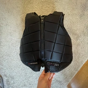 Svart ridväst  från Racesafe - Säljer en svart ridväst från Racesafe med dragkedja framtill. Västen är svart och man kan spänna in den på sidorna! Den har bra skick! Säljer pg att den är för liten annars är den jätte bekväm att rida i ock flexibel! Nypris 3400 
