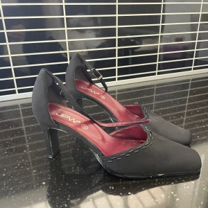 Svarta klackskor från JPW - Eleganta svarta klackskor från JPW med spetsig tå och justerbart spänne runt ankeln. Skorna har en stilren design med dekorativa sömmar längs kanten och en bekväm röd innersula.