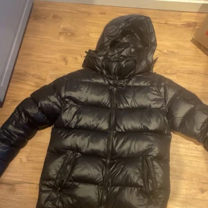 Svart dunjacka från Moncler - Snygg svart dunjacka från Moncler med huva och dragkedja. Jackan har en quiltad design och är perfekt för kalla dagar. Insidan är ljusgrå och har en unik etikett med serietidningsmotiv.