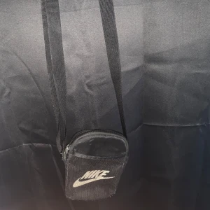 Svart axelväska från Nike - Säljer en smidig svart axelväska från Nike med justerbar rem och dragkedja. Väskan har en röd insida och är perfekt för att bära med sig småsaker. Den har en klassisk Nike-logga på framsidan.