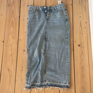 Jeanskjol med rå kant - Säljer en cool jeanskjol med rå kant och knappar framtill. Kjolen har en klassisk blå denimfärg och en rak passform som ger en avslappnad look. Perfekt för en casual stil! Har en liten fläck framtill som går att tvätta bort.