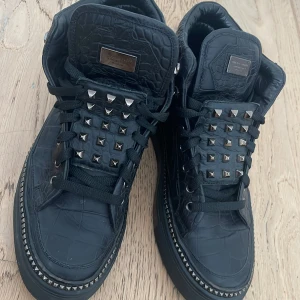 Svarta sneakers från Philipp Plein - Snygga svarta sneakers från Philipp Plein med krokodilpräglat mönster och coola nitar på ovansidan. Skorna har snörning och är tillverkade i Italien. Perfekta för dig som vill ha en unik stil. PRISET KAN ALLTID DISKUTERAS