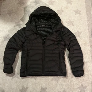 Svart pufferjacka från J.Lindeberg - Säljer en stilren svart pufferjacka från J.Lindeberg. Jackan har en praktisk huva och dragkedja framtill. Perfekt för kyligare dagar med sin quiltade design som håller värmen. Passar bra till både jeans och mer avslappnade outfits. Funkar nu till våran då den inte är lika tjock som en vinterjacka.  Mycket bra skick och inga defekter 