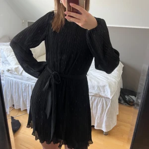 Svart plisserad klänning med knytband - Snygg svart plisserad klänning med långa ärmar och ett knytband i midjan. Klänningen har en kort längd och ger en stilren siluett. Den är endast använd ett fåtal gånger🙌🏻🤩