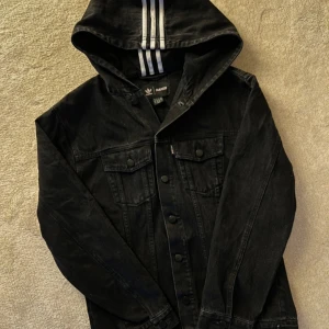 Svart jeansjacka AdidasxNaked - Säljer en svart jeansjacka från Adidas med huva och de klassiska tre ränderna i vitt. Jackan har knappar framtill och två bröstfickor med lock. Perfekt för en sportig och avslappnad stil.