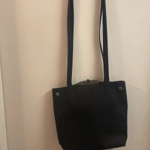 Svart axelväska - Snygg svart axelväska med långa remmar och en stilren design. Perfekt för att bära med sig det nödvändigaste. Väskan har en enkel stängning upptill och är lätt att matcha med olika outfits.