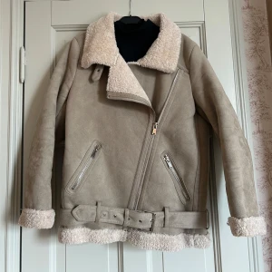 Beige bikerjacka pilotjacka från Zara - Snygg beige jacka i fejk mocka från Zara med mjukt foder och detaljer i fårskinnslook. Jackan har en asymmetrisk dragkedja, bälte i midjan och två snedställda fickor med dragkedjor. Använd och har därför en väldigt liten fläck på ena armen - se bild 3.