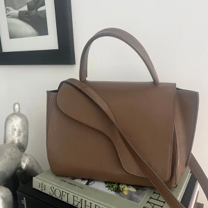 Brun ATP handväska i skinn - Denna eleganta bruna läderväska är både tidlös och sofistikerad. Trots sin mindre storlek rymmer den allt du behöver, perfekt för en stilfull vardag eller speciella tillfällen. Dess design utstrålar klassisk elegans och är en investering som varar.