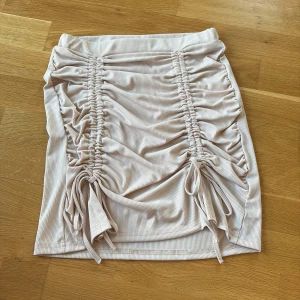 Kjol beige  - En beige ribbad kjol med detaljer framtill som man kan justera längden på och knyta själv, resår band i midjan, aldrig använd 