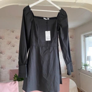 Svart klänning från NA-KD - Elegant svart klänning från NA-KD med långa ärmar och fyrkantig urringning. Klänningen har knappar vid ärmsluten och en smickrande passform som passar perfekt för olika tillfällen.