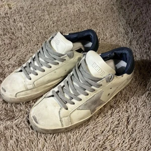 Golden goose skor - Snygga vita sneakers från Golden Goose med en grå stjärna på sidan. Skorna har grå snörning och en mörkblå häl. Perfekta för en avslappnad stil.