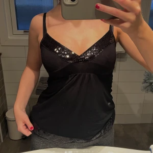 Svart paljettlinne - Snyggt svart linne med paljettdetaljer upptill och längs nederkanten. Linnet har smala axelband och en v-ringad design som ger en elegant look. Perfekt för en kväll ute eller festliga tillfällen.