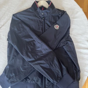 Svart vindjacka från Moncler - Snygg svart vindjacka från Moncler med dragkedja och ribbad krage. Jackan har ett broderat Moncler-märke på ärmen och stängs med en knapp vid halsen. Perfekt för en stilren look. Köpt från careofcarl, pris kan självklart diskuteras😊