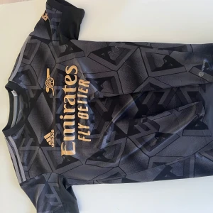 Svart Arsenal tröja  från Adidas - Säljer en svart fotbollströja från Adidas med guldiga detaljer och ett geometriskt mönster. Tröjan har korta ärmar och en rund halsringning. Perfekt för fotbollsfans som vill ha en stilren look på planen. Använd fåtal gånger