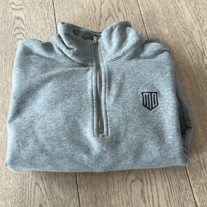 Grå hoodie från Maya Delorez - Säljer en stilren grå hoodie från Maya Delorez med halv dragkedja och broderad logga på bröstet. Perfekt för en avslappnad look. Designad i Sverige. Säljer då den e för liten