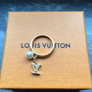 Louis Vuitton Guldring med pärla och Louis Vuitton-berlock - Elegant guldring från Louis Vuitton med en hängande pärla och den ikoniska LV-berlocken. Perfekt för att ge en lyxig touch till din stil. Kommer i originalförpackning med en snygg förvaringspåse. 19mm