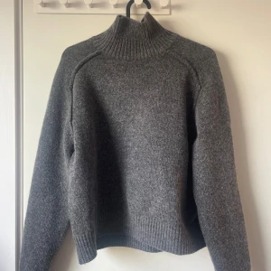 Grå stickad polotröja - Säljer en mysig grå stickad polotröja med ribbade detaljer vid krage, ärmslut och nederkant. Perfekt för kyliga dagar och ger en stilren look. Passar bra till jeans eller kjol.