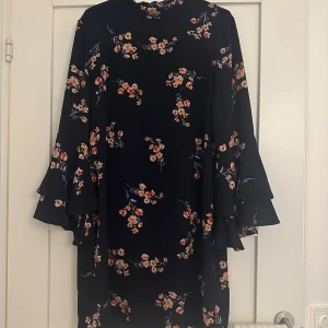 Blommig klänning med volangärmar - Snygg mörkblå klänning med blommönster i orange och vitt. Klänningen har långa volangärmar och en lös passform. Perfekt för en stilfull look.