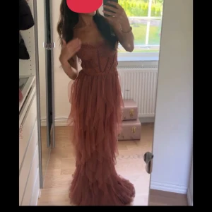 Brun långklänning med volanger - Elegant brun långklänning med volanger och spetsdetaljer. Klänningen är axelbandslös och har en vacker, flytande design som ger en romantisk känsla.