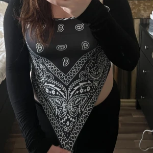 Svart och vit bandanamönstrad topp - Snygg svart och vit topp med bandanamönster. Toppen har en unik design med en triangulär framsida och långa ärmar. Perfekt för en trendig look.