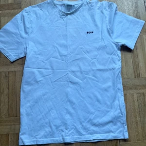 Vit t-shirt från Boss - Säljer en klassisk vit t-shirt från Boss med korta ärmar och rund hals. T-shirten har en enkel design med Boss-loggan tryckt på bröstet. Perfekt för en stilren och avslappnad look.