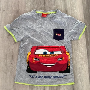Grå t-shirt med Blixten McQueen från Disney Pixar - Cool grå t-shirt med Blixten McQueen från Disney Pixar Cars. Framsidan har en stor bild av Blixten och texten 'Let's see what you got!'. Baksidan har texten 'Way ahead of you!' och en bild av Blixtens bakdel. *Barn size