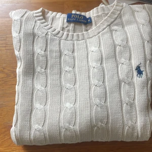 Vit stickad tröja från Ralph Lauren - Säljer en stilren vit stickad tröja från Ralph Lauren med kabelstickat mönster och den klassiska blå loggan på bröstet. Tröjan har långa ärmar och rund halsringning. Perfekt för en elegant och avslappnad look.
