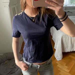 Mörkblå kortärmad topp med guldknappar - Säljer en mörkblå topp från Zara med korta ärmar och dekorativa knappar på axeln. Perfekt för en stilren look.