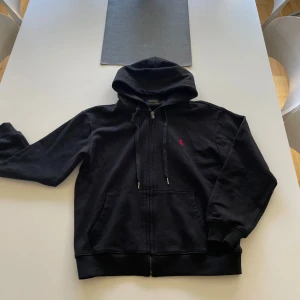 Svart Zip hoodie från Polo Ralph Lauren - Säljer en svart hoodie från Polo Ralph Lauren med dragkedja och en broderad röd logotyp på bröstet. Tröjan har en justerbar huva och fickor framtill. Perfekt för en avslappnad stil.