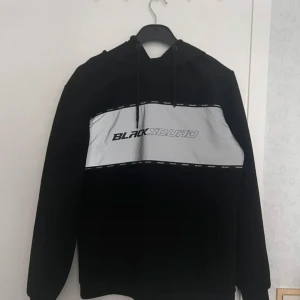 Svart hoodie med reflexdetalj - en svart hoodie från BLACKSQUAD med stor reflexpanel och logga över bröstet. Slim fit. Använd en gång! Har några streck på sig som jag inte tror tillhör designen? 😅Axelbredd 48cm, längd 72cm.