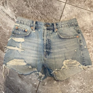 Ljusblå jeansshorts med slitningar från Zara - Säljer ett par ljusblå jeansshorts från Zara med råa kanter och tydliga slitningar framtill. Klassisk femficksmodell med hög midja och knappgylf. Perfekta för en avslappnad stil.