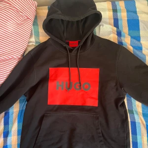 Svart hoodie från Hugo Boss med röd logga - Säljer en svart hoodie från Hugo Boss med stor röd logga och texten HUGO på bröstet. Tröjan har huva med dragsko och en stor magficka. Perfekt för en stilren och avslappnad look.