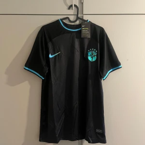 Svart Brasilien fotbollströja från Nike - Säljer en svart Brasilien fotbollströja från Nike med turkosa detaljer och CBF-emblem på bröstet. Tröjan har korta ärmar och rund hals med turkos kant. Perfekt för dig som vill sticka ut på planen eller heja på Brasilien.