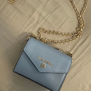 Ljusblå axelväska från Michael Kors - Säljer en stilren ljusblå axelväska från Michael Kors med guldfärgad kedja och logga framtill. Väskan har en elegant, fyrkantig form och är perfekt för att bära det viktigaste. Enkel och snygg design som passar till många tillfällen. Kan även användas som plånbok