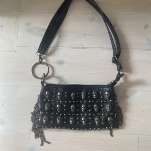 Svart axelväska med dödskalle-detaljer - Cool svart axelväska med silverfärgade dödskallar och nitar på framsidan. Väskan har kedjedetaljer på sidorna, dragkedja upptill och en bred axelrem med stora metallringar. Perfekt för dig som vill sticka ut med en edgy accessoar.