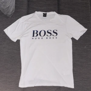 Vit t-shirt från Hugo Boss - Vit t-shirt från Hugo Boss med stor BOSS-logga tryckt i mörkblått på bröstet. Klassisk rund halsringning och korta ärmar. Perfekt för en clean och stilren look.