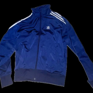 Blå Adidas track jacket med vita ränder - Klassisk blå track jacket från Adidas med vita ränder längs axlarna och dragkedja framtill. Kan mådde användas som en vardags plagg men även som tränings tröja ny pris ca 800kr