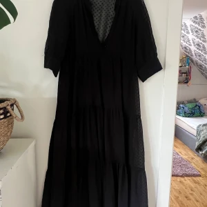 Svart prickig långklänning från Zara - Svart långklänning från Zara med diskret prickigt mönster och v-ringning. Klänningen har korta ärmar och volangdetaljer i kjolen. Tunt, lätt material som ger en luftig känsla. Perfekt för dig som gillar en flowy och stilren look.
