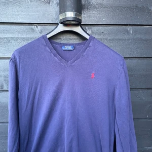 Blå v-ringad tröja från Polo Ralph Lauren - Säljer en klassisk blå v-ringad tröja från Polo Ralph Lauren med röd broderad logga på bröstet. Tröjan har långa ärmar och är tillverkad i mjukt material som passar perfekt till många tillfällen. Storlek L.