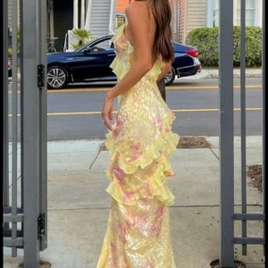 Ljusblommig långklänning med volanger - Romantisk långklänning i ljusa toner med blommigt mönster och volangdetaljer. Klänningen har smala axelband och v-ringad halsringning. Perfekt för sommarens speciella tillfällen.