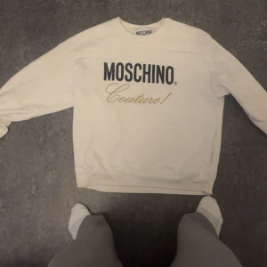 Vit sweatshirt från Moschino Couture - Säljer en vit sweatshirt från Moschino Couture med tryckt logga i svart och guld på bröstet. Tröjan har rund halsringning och långa ärmar. Perfekt för en stilren och trendig look.