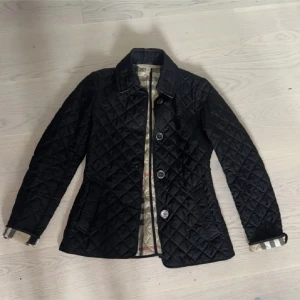 Burberry jacka  - Snygg svart quiltad jacka från Burberry med klassiskt rutigt foder. Jackan har lång ärm, knappar framtill och två fickor. Perfekt för höst och vår, med en tidlös design och fina detaljer.