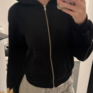 Svart hoodie med dragkedja - Svart hoodie med dragkedja framtill och huva. Modellen har normal passform och två fickor på magen. Perfekt för en avslappnad stil och enkel att matcha med andra plagg.