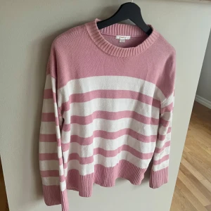 Rosa och vit randig stickad tröja - Superfin stickad tröja i rosa och vitt med breda ränder. Tröjan har rund halsringning, långa ärmar och ribbade muddar vid ärmslut och nederkant. Perfekt för en mysig och avslappnad stil.