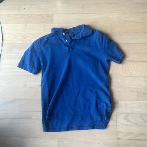 Blå pikétröja från Polo Ralph Lauren - Säljer en klassisk blå pikétröja från Polo Ralph Lauren med röd broderad logga på bröstet. Tröjan har korta ärmar, krage och knappar vid halsen. Perfekt för en stilren och avslappnad look.