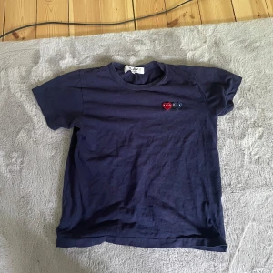 Mörkblå t-shirt från Comme des Garçons Play - Säljer en mörkblå t-shirt från Comme des Garçons Play med två broderade hjärtan i rött och svart på bröstet. Klassisk passform och korta ärmar. Perfekt för dig som gillar stilrena plagg med en twist.