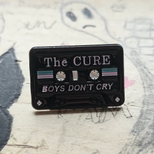 The Cure - Boys don't cry PIN - En svart pin i form av en kassett med texten 'The Cure Boys Don't Cry' i vitt och lila. Detaljer i turkos och lila på framsidan.