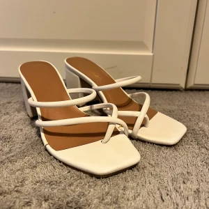 Vita sandaler med klack och remmar - Säljer ett par trendiga vita sandaler med fyrkantig tå och bred klack. Skorna har flera tunna remmar över foten och öppen tå, vilket ger en modern och stilren look. Perfekta studenyklackar
